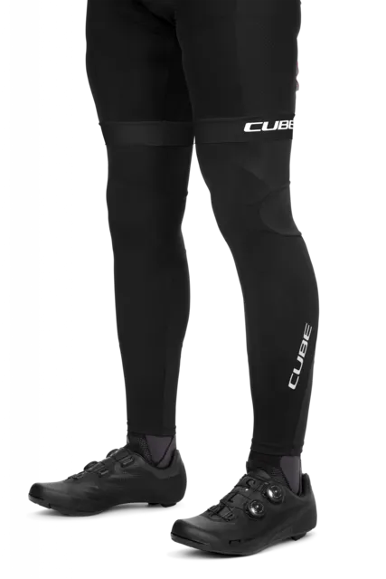 Eine Person trägt CUBE BLACKLINE Beinlinge und schwarze Fahrradschuhe, wobei "CUBE" auf den Beinlingen und Schienbeinen zu sehen ist. Die Unterschenkel und Füße sind vor einem weißen Hintergrund abgebildet.