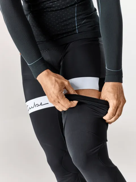 Eine Person in schwarzer CUBE BLACKLINE Beinlinge zieht ihre eng anliegende Hose hoch, so dass weiße Streifen und ein Teil des Oberschenkels sichtbar werden. Die Kleidung trägt einen Schriftzug und ein gepunktetes Muster auf dem Oberteil., Ansicht 4