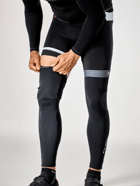 Eine Person in schwarzer Sportkleidung und Fahrradschuhen zieht die CUBE BLACKLINE Beinlinge über die Oberschenkel, wobei die reflektierenden Bänder oberhalb der Knie sichtbar sind; nur der Körper von den Schultern bis zu den Füßen ist zu sehen., Ansicht 5