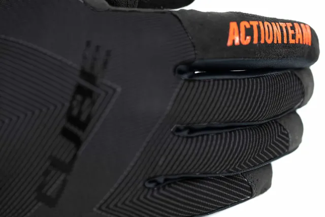 Eine Nahaufnahme des CUBE Handschuhe Performance langfinger X Actionteam Handschuhs in schwarz, mit fein strukturierten Linien und "ACTIONTEAM" in kräftigem Orange auf einem Finger, wie er von Mr. Bike Lichtenfels getragen wird., Ansicht 4