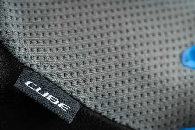 Nahaufnahme des grau strukturierten CUBE Handschuhe langfinger X NF Handschuhs, der ein kleines schwarzes Etikett mit "CUBE" in Weiß, ein gitterartiges Muster und einen Teil eines blauen Logos aufweist - eine Qualität, die an die Ausrüstung von Mr. Bike Lichtenfels erinnert., Ansicht 3