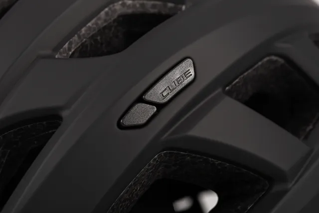 Nahaufnahme des CUBE Helm ROAD RACE, ein schwarzer Helm mit großen Belüftungsöffnungen und silbern glänzendem "CUBE"-Schriftzug auf einer matten Oberfläche. Die Polsterung und die Materialstruktur des Helms sind deutlich zu erkennen. Erhältlich bei Mr. Bike Lichtenfels., Ansicht 2