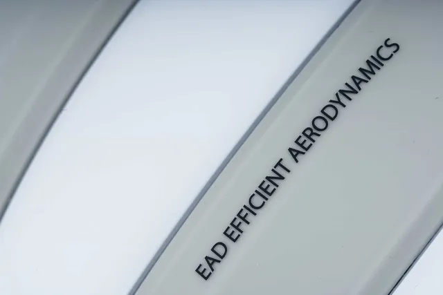 Nahaufnahme einer weißen Fläche mit geschwungenen Linien und schwarzem Text mit der Aufschrift "EAD EFFICIENT AERODYNAMICS", die sich wahrscheinlich auf den CUBE Helm HERON bezieht. Der Anfang des ersten Wortes ist abgeschnitten, was auf "Mr. Bike Lichtenfels" im Design oder Branding hinweist., Ansicht 4