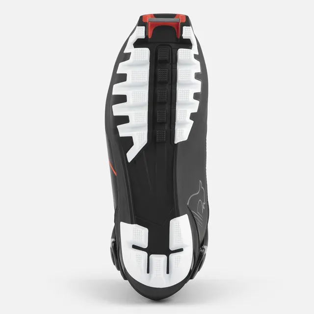Die Abbildung zeigt die Sohle des ROSSIGNOL X-6 SC, eines schwarzen Sportschuhs mit weißen und roten Akzenten. Das strukturierte Profil und die erhabenen Bereiche sorgen für Grip und Stabilität, während im Fersenbereich ein schwacher Schriftzug zu sehen ist.