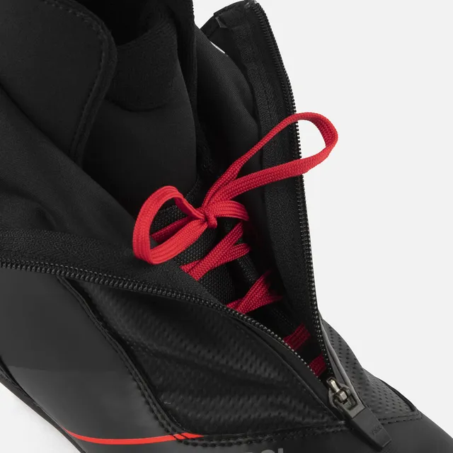 Nahaufnahme des ROSSIGNOL X-6 SKATE, eines schwarzen Sportschuhs mit roten Schnürsenkeln, der teilweise unter einer halb geöffneten schwarzen Hülle sichtbar ist, die das Innenfutter und einen Teil der Zunge offenbart.