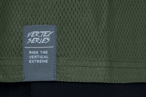 Nahaufnahme des strukturierten olivgrünen Gewebes des CUBE VERTEX Rundhalstrikot TM langarm, mit grauem Etikett: VERTEX SERIE. FAHREN SIE DIE VERTIKALEN EXTREME. Das CUBE VERTEX-Label ist in der Nähe des Saums aufgenäht.