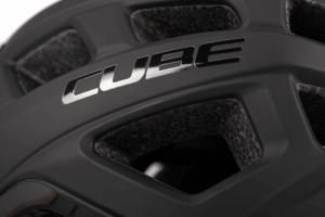 Nahaufnahme des CUBE Helm ROAD RACE, ein schwarzer Helm mit großen Belüftungsöffnungen und silbern glänzendem "CUBE"-Schriftzug auf einer matten Oberfläche. Die Polsterung und die Materialstruktur des Helms sind deutlich zu erkennen. Erhältlich bei Mr. Bike Lichtenfels.
